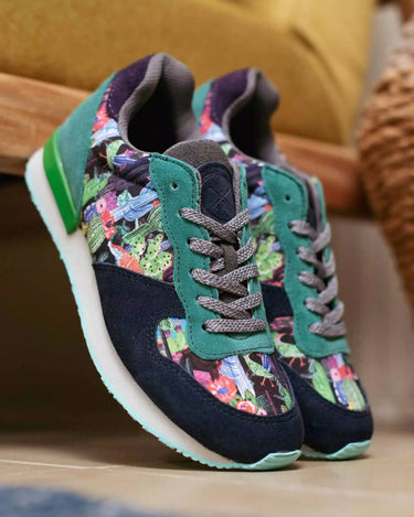 A pair of Inkkas joggers sneakers with a dark navy suede base, teal green overlays, and a colorful floral pattern on the side panels, resting on a wooden surface in a home setting.