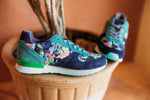 A pair of Inkkas jogger sneakers displayed on a terracotta planter. The sneakers feature a navy blue suede base with a colorful, busy abstract pattern across the toe box and side panels, teal suede overlays, a bright green heel accent, and a white midsole with a light blue outsole.