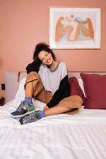 A woman sits on a bed wearing a color-blocked knit sweater, brown shorts, and colorful sneakers, with a pink wall and framed art behind her.
