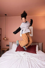 A woman jumps excitedly on a bed in a bedroom with dusty pink walls. She is wearing a light gray knit sweater over a black long-sleeve shirt, loose tan pants, and colorful sneakers.