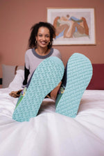 A smiling woman sits on a white bed with her legs raised, showing the textured light blue soles of her sneakers. The room has a dusty rose wall with a framed abstract painting in the background.