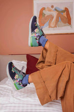 A person reclining on a bed wearing colorful sneakers with floral graphics, blue socks, and wide-leg brown pants, with a framed abstract painting on a peach wall in the background.