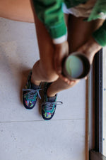 A person sitting on a light-colored floor holds a metal cup while wearing colorful embroidered sneakers with green tops and gray laces.