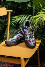 A pair of colorful sneakers with a vibrant graffiti-style print, displayed on a yellow wooden chair surrounded by lush green tropical foliage.
