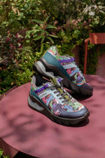 A pair of colorful, highly patterned athletic sneakers displayed on a dusty rose table outdoors with green foliage in the background.