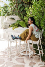 A woman sits on a white metal patio barstool outdoors, smiling and looking to the side. She is wearing a light gray sleeveless top with a red graphic, olive green pants, and colorful sneakers.