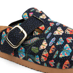 A close-up of a clog-style shoe featuring a navy blue fabric upper with a colorful butterfly and leaf print, a matching fabric strap with a gold-tone buckle, and a brown sole.