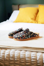 A pair of slip-on clogs with a colorful floral pattern on a dark background, featuring a brown sole and a gold buckle, resting on a bed with yellow pillows.