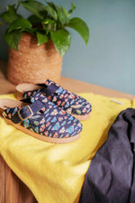 A pair of slip-on clogs with a colorful fish pattern on a navy blue background, styled on a yellow blanket next to a dark gray fabric, with a potted green plant in the background.