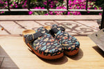 A pair of clogs with a colorful butterfly pattern on a black background, resting on a wooden deck with a blurred background of flowers and a metal railing.