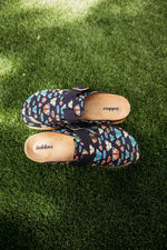 A pair of Inkkas slip-on clogs with a colorful geometric pattern on a dark navy base, placed on green grass.