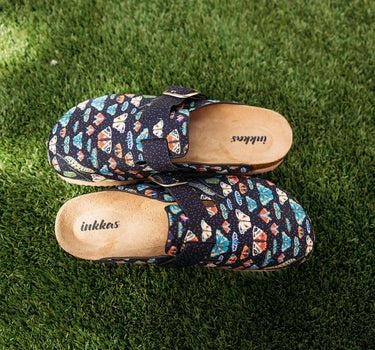 A pair of Inkkas slip-on clogs with a colorful geometric pattern on a dark navy base, placed on green grass.