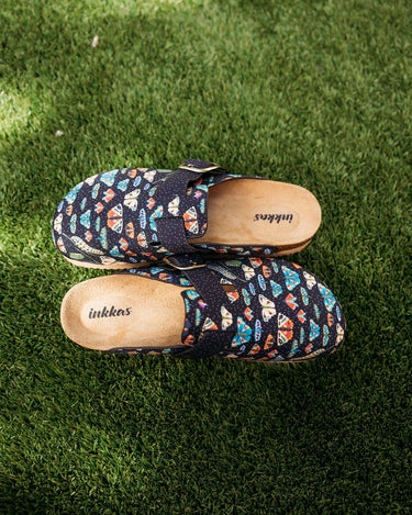 A pair of Inkkas slip-on clogs with a colorful geometric pattern on a dark navy base, placed on green grass.