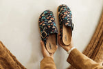 A pair of black slip-on clogs with a colorful butterfly pattern and gold buckle, held by hands wearing brown suede sleeves against a white background.