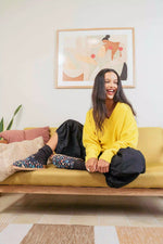 A woman sits smiling on a mustard yellow sofa in a bright living room, wearing a yellow sweater, black pants, and patterned socks. A framed abstract painting hangs on the wall behind her.