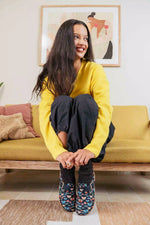 A woman sitting on a mustard yellow couch, wearing a bright yellow sweater, dark pants, Inkkas clogs and chunky socks. She is smiling and looking to the side.