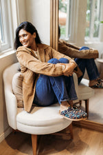 A woman sits in a cream-colored armchair in a bright room, wearing a tan suede jacket, blue jeans, and patterned slip-on shoes. Her reflection is visible in a mirror behind her.