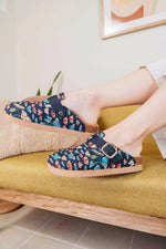 A person wearing a pair of slip-on clog-style slippers with a navy blue floral pattern and a gold-tone buckle, sitting on a mustard-yellow upholstered bench.