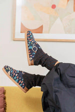 A person reclining indoors with legs raised, wearing dark patterned slip-on shoes with a buckle strap and dark knit socks, against a light wall with framed abstract art and a mustard-yellow surface below.