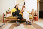 Two women laughing and sitting on a yellow-green sofa in a modern living room. The woman on the left wears a bright yellow sweatshirt, black pants, and colorful patterned slip-on shoes, while the woman on the right wears a white sleeveless top and white pants with matching colorful slip-on shoes.
