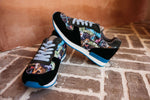 A pair of black low-top sneakers with colorful graffiti-style printed panels, gray laces, and blue accents, resting on a brick floor against a terracotta wall.