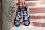 A person holding a pair of low-top sneakers with a colorful illustrated pattern on the toe boxes and sides, black mesh uppers, and light blue laces. The person is wearing khaki cargo pants, with blurred terracotta steps in the background.