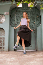 A woman posing in front of a teal metal gate, wearing a lavender sleeveless top, a black high-low skirt, and gray athletic sneakers.