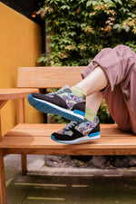A person sitting on a wooden bench outdoors wearing black low-top sneakers with a colorful floral pattern on the sides, bright blue soles, and light green socks.