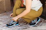 A person crouching down to tie the laces of black and blue sneakers with colorful floral patterns, wearing olive green pants and a white t-shirt on a stone patio.