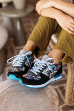 A person sitting indoors wearing black sneakers with a colorful printed pattern on the sides, white laces, and bright blue soles, seated on a wooden stool.