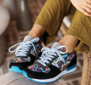 A person sitting indoors wearing black sneakers with a colorful printed pattern on the sides, white laces, and bright blue soles, seated on a wooden stool.