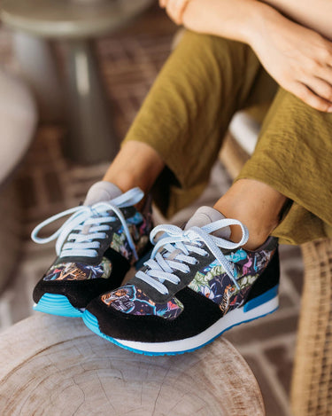 A person sitting indoors wearing black sneakers with a colorful printed pattern on the sides, white laces, and bright blue soles, seated on a wooden stool.