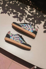 A pair of low-top sneakers with a colorful graphic pattern on the uppers, black toe and heel caps, and gum soles, resting on a light-colored outdoor table under leafy shadows.