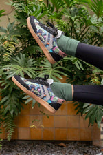 A person wearing colorful patterned skate shoes, green crew socks, and black leggings, posed with their legs raised against a background of green plants and a tiled wall.