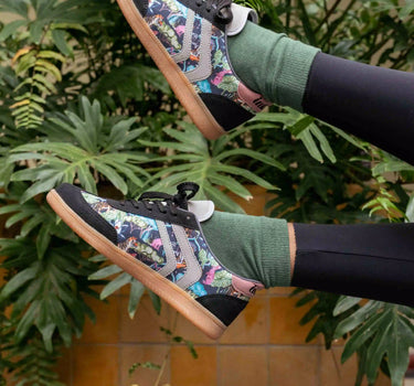A person wearing colorful patterned skate shoes, green crew socks, and black leggings, posed with their legs raised against a background of green plants and a tiled wall.