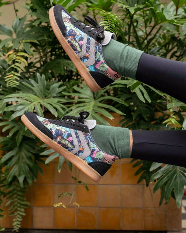 A person wearing colorful patterned skate shoes, green crew socks, and black leggings, posed with their legs raised against a background of green plants and a tiled wall.