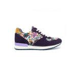 A single low-top sneaker shown in profile against a white background. The shoe features dark purple suede panels, a multicolor floral print overlay on the quarter and toe sections, a yellow heel tab, a light purple midsole accent, and a white midsole.