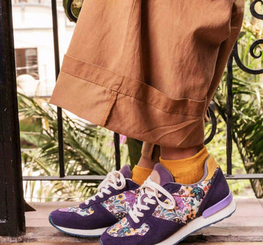 A person wearing purple sneakers with a colorful floral pattern on the sides, styled with mustard yellow socks and brown wide-leg trousers, standing on a wooden deck.