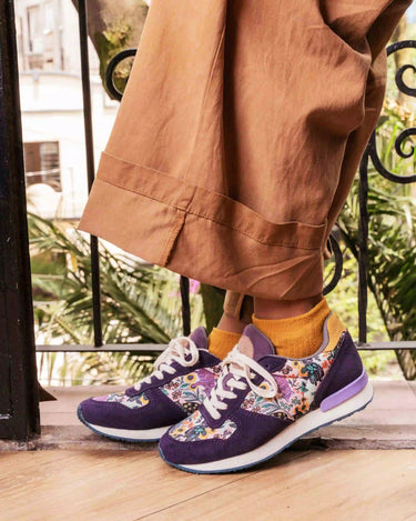 A person wearing purple sneakers with a colorful floral pattern on the sides, styled with mustard yellow socks and brown wide-leg trousers, standing on a wooden deck.