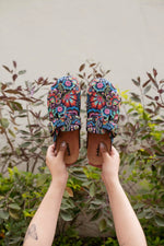 A pair of colorful embroidered clogs held up by two hands against a background of green plants and a beige wall. The clogs feature vibrant floral embroidery in blue, red, pink, and green on a black base, with brown leather soles.