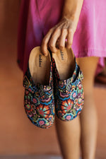 A person in a pink dress holds a pair of colorful embroidered slip-on shoes with a floral pattern. The insoles show the brand name Inkkas.