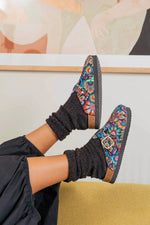 A person wearing colorful floral slip-on mule shoes with black knit socks, shown from the knees down in a styled indoor setting.