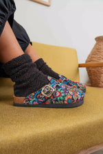 A person wearing colorful floral slip-on clogs with a gold buckle and black knit socks, sitting on a mustard yellow upholstered chair.
